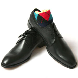 Foto von zwei schwarzen Derby Glattleder Herrenschuhen. Der rechte Schuh steht auf dem anderen und ein buntes Sockenpaar schaut heraus. Modell Stil Statement