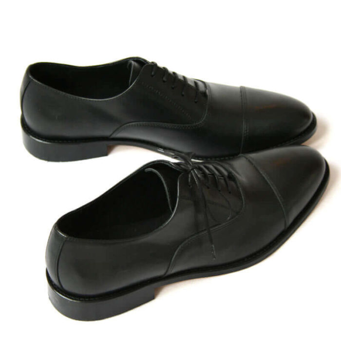 Foto von zwei schwarzen Oxford Glattleder Herrenschuhen mit Zehenkappe. Beide nach rechts zeigend. Einer quer stehend. Die Schuhspitzen berühren sich. Modell Oxford Pro