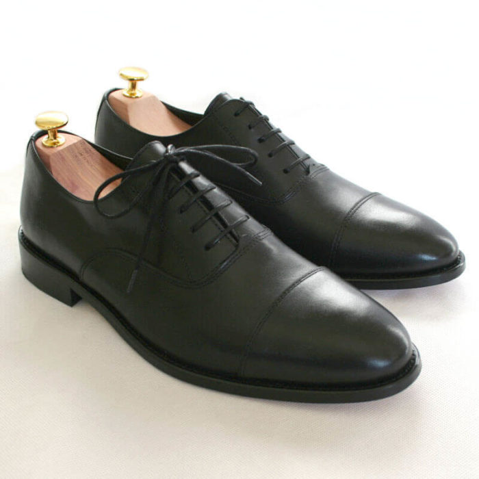 Foto von zwei schwarzen Oxford Glattleder Herrenschuhen mit Zehenkappe. Beide zeigen nach rechts unten und sind mit Schuhspannern mit Goldknöpfen versehen. Modell Oxford Pro