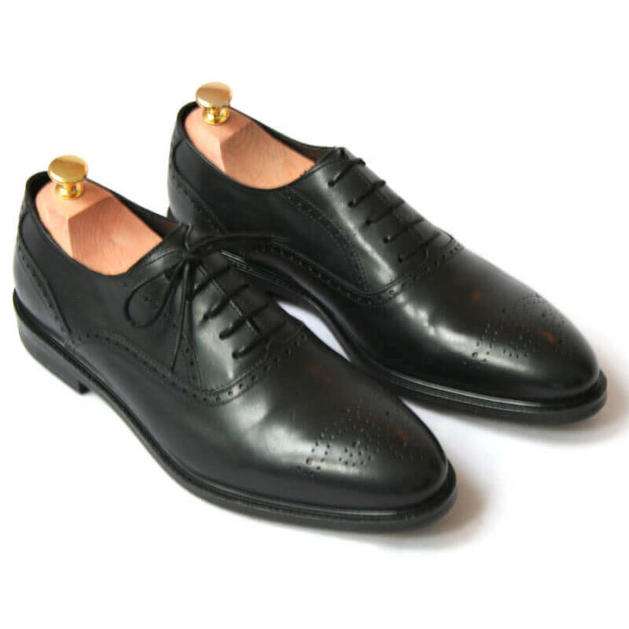 Foto von zwei schwarzen Oxford Glattleder Herrenschuhen mit Verzierung. Beide zeigen nach rechts unten und sind mit Schuhspannern mit Goldknöpfen versehen. Modell Oxford Plus