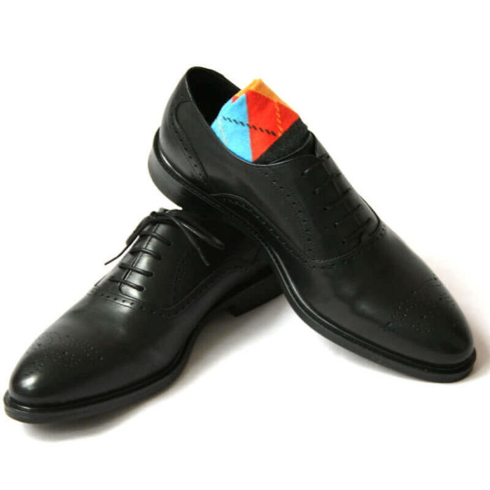 Foto von zwei schwarzen Oxford Glattleder Herrenschuhen mit Verrzierung. Der rechte Schuh steht auf dem anderen und ein buntes Sockenpaar schaut heraus. Modell Oxford Plus