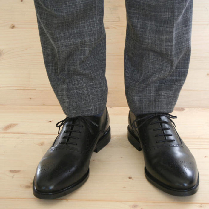Foto von Herrenbeinen im grauen Anzug mit zwei schwarzen Oxford Glattleder Herrenschuhen mit Verzierung. Modell Oxford Plus