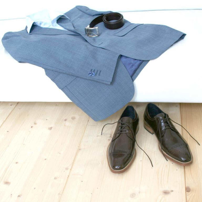 Foto-Zwei braune Herrenschuhe mit Teilperforation und gemusterter Zehenkappe vor weißer Couch auf der ein hellblauer Anzug mit Hemd und Gürtel liegt-8