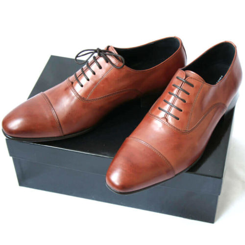 Foto von zwei braunen Oxford Glattleder Herrenschuhen mit Zehenkappe auf schwarzem Schuhkarton. Modell Oxford first
