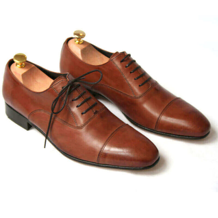 Foto von zwei braunen Oxford Glattleder Herrenschuhen mit Zehenkappe. Beide zeigen nach rechts unten und sind mit Schuhspannern mit Goldknöpfen versehen. Modell Oxford first