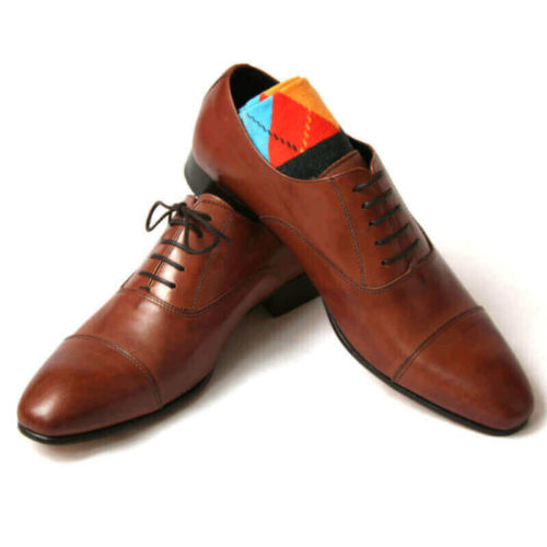 Foto von zwei braunen Oxford Glattleder Herrenschuhen mit Zehenkappe. Der rechte Schuh steht auf dem anderen und ein buntes Sockenpaar schaut heraus. Modell Oxford first
