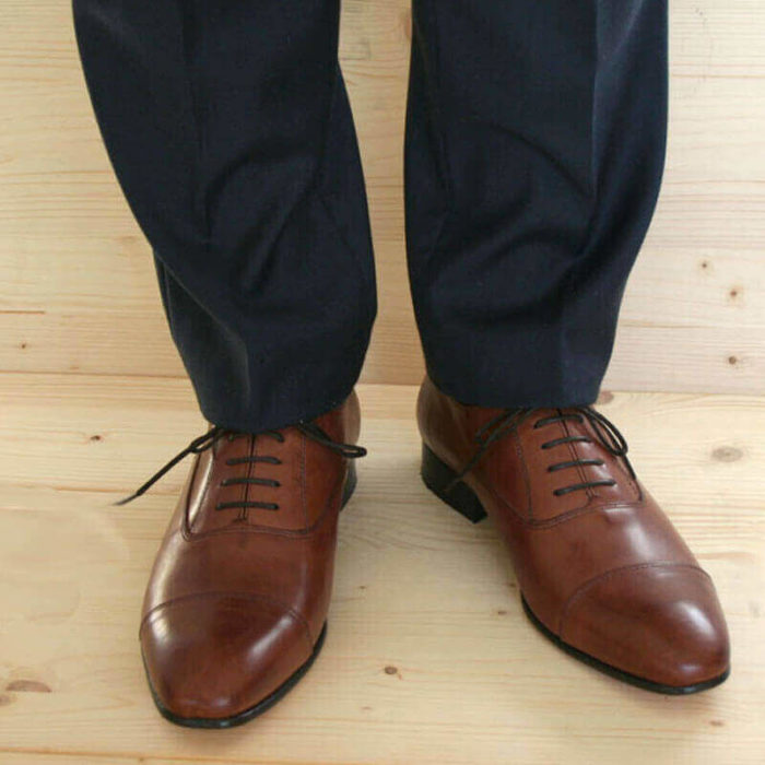 Foto von Herrenbeinen im grauen Anzug mit zwei braunen Oxford Glattleder Herrenschuhen mit Zehenkappe. Modell Oxford first