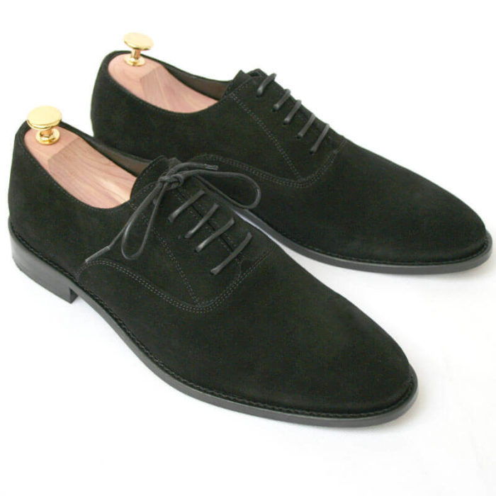 Foto von zwei schwarzen Wildleder Oxford Herrenschuhen nach schräg unten rechts zeigend mit Goldkopf-Schuhspannern. Modell Herren Oxford