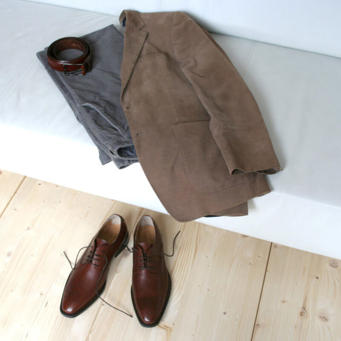 Foto-Nussbraune Business Herrenschuhe_auf hellem Holzfußboden abgestellt, dahinter graue Hose, hellbraunes Jacket und nussbrauner Gürtel