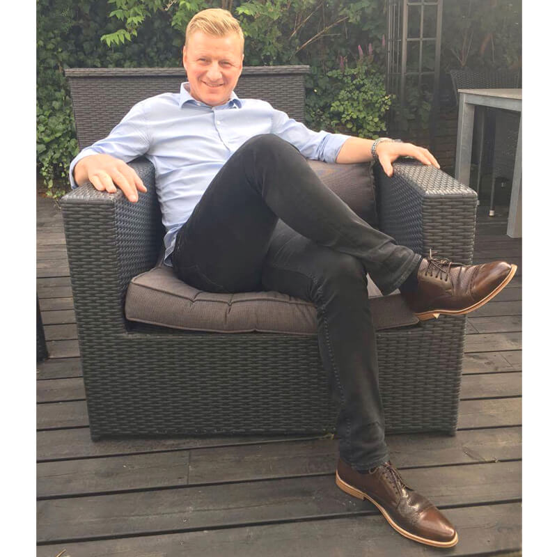 Foto: Mann sitzend auf Terrasse in einem Outdoor-Sessel mit hellblauem Oberhemd und schwarzer Hose dazu braune Herrenschuhe Komfortabler Klassiker